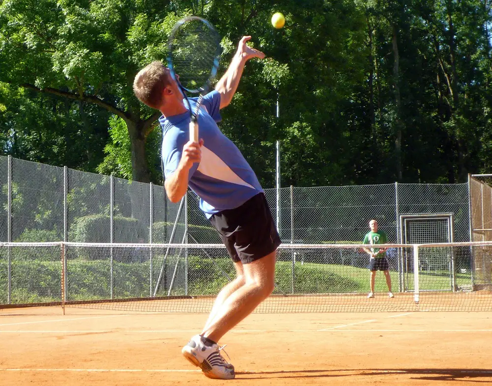 2 jeunes jouant au tennis . Le premier est en train de servir au premier plan et l'autre est en position de réception de l'autre côté du court