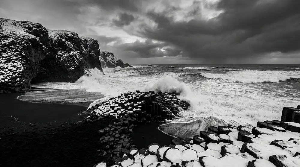 Plage de sable noir de Reynisfjara avec colonnes basaltiques, vagues de l'Atlantique et ciel dramatique en hiver