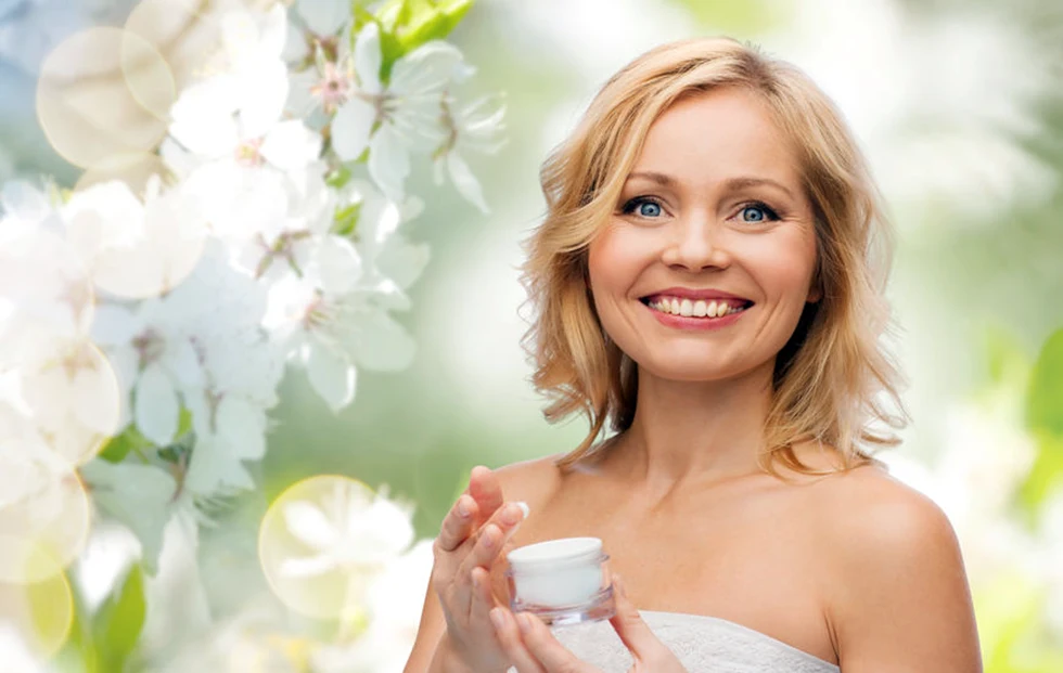 Femme blonde souriante et rayonnante avec un arrière plan naturel , tenant un pot de crème de soin du visage